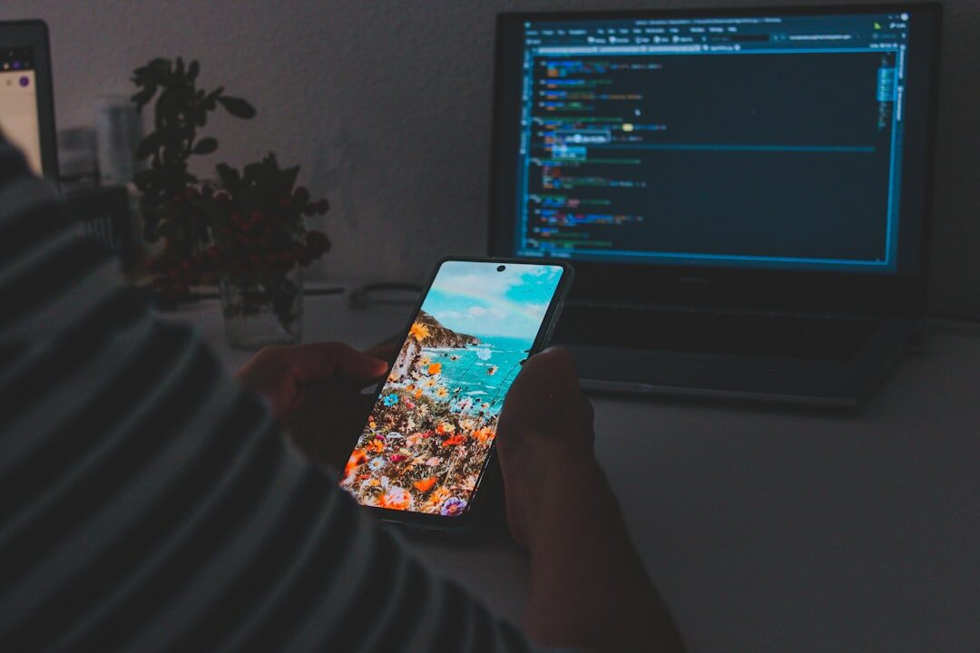 Young developer holding a smartphone while coding on a laptop